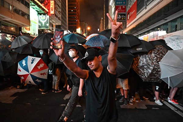 A protester shows the Hong-Kong emblem on his mobile phone and flashes a victory sign as he stands in front of other protesters carrying umbrellas to face the police outside the West Kowloon railway station during a demonstration against a proposed extradition bill in Hong Kong on July 7, 2019. - Tens of thousands of anti-government protesters rallied outside a controversial train station linking the territory to the Chinese mainland on July 7, the latest mass show of anger as activists try to keep pressure on the city's pro-Beijing leaders. (Photo by HECTOR RETAMAL / AFP) 