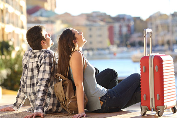 Side view of a couple of 2 tourists with a suitcase sitting relaxing and enjoying vacations in a colorful promenade. Tourism concept Fotolia