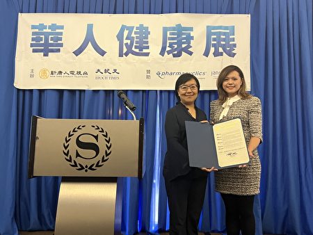 纽约州众议员金兑锡（Ron Kim）向新唐人健康展颁发褒奖，由他的社区联络主任冯子馨（右）代为向新唐人电视台市场总监Joyce Jiao（左）颁发。