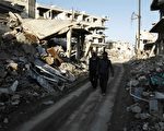 2016年2月27日，敘利亞停火協議幾小時後，西部的塞萊米耶（Salamiyeh）市又發生炸彈攻擊，造成2死4傷。本圖為24日，2名敘利亞人走在大馬士革東部郊區的街道上，道路兩旁盡是飽受戰火摧殘的斷垣殘壁和廢墟。（ABDULMONAM EASSA/AFP/Getty Images）