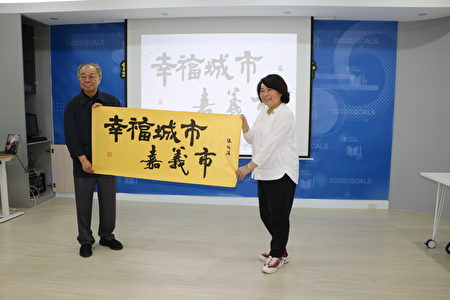  淡江大学张炳煌教授送字画给市长黄敏惠，与背后投影的，以平板挥毫的书法字几乎毫无差距。