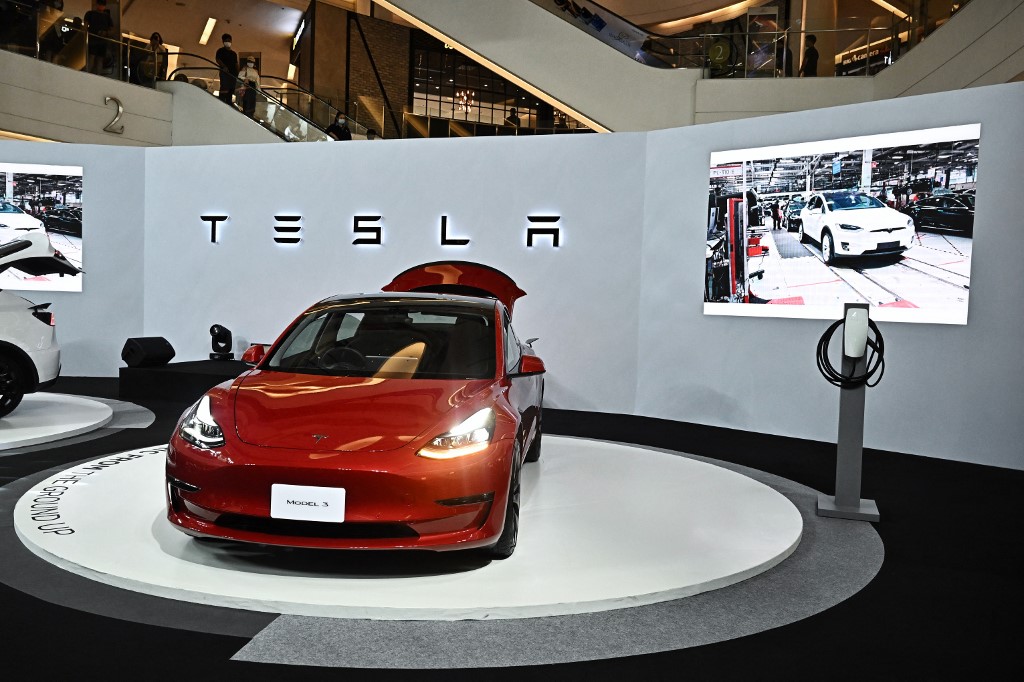 A red Tesla Model 3 is showcased on a circular platform at a display booth with large screens and a white backdrop, highlighting Tesla's electric vehicles amidst market competition and recent product simplifications.