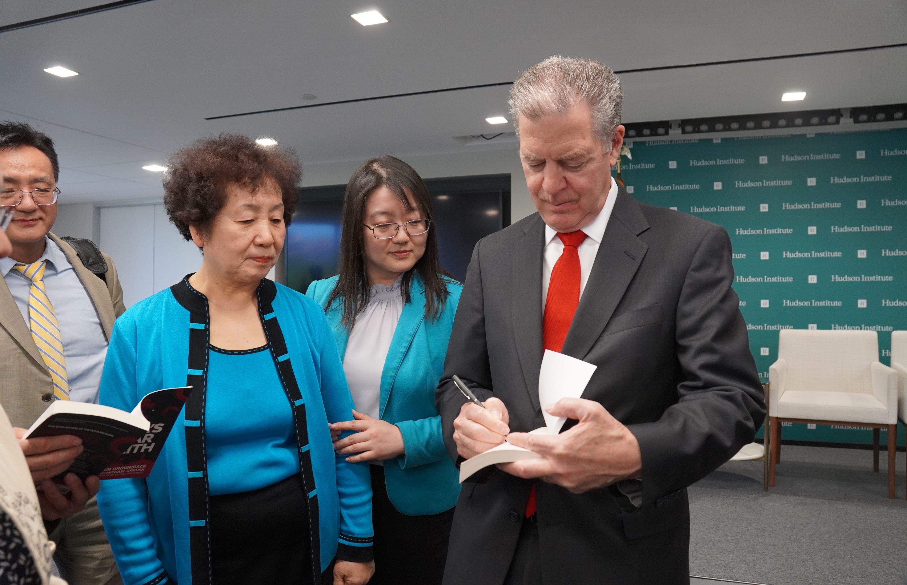 2026年5月12日,美國前宗教自由大使薩姆‧布朗巴克(Sam Brownback)在華府智庫哈德遜研究所發布新書《中共對信仰的戰爭》(China’s war on Faith)。圖為原遼寧省大連法輪功學員王春彥(前排穿藍色衣服者)在活動上請布朗巴克大使(右)在其新書上簽名。(李辰/大紀元)
